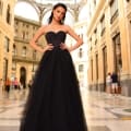 A woman stands confidently in an opulent, sunlit indoor setting, wearing NC1019, a strapless black gown with a full tulle skirt from Sunshine Coast Dress Shop. Her hands are on her hips, and she has long, dark hair. The background features a high, arched glass ceiling and grand architectural details. Angels Formal Wear