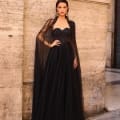 In front of a textured stone wall, a woman confidently poses in an NC1019 gown. The elegant black dress is adorned with intricate details and features a flowing sheer cape. She completes her stylish look with large statement earrings and places one hand on her hip, embodying the essence of the NC1019 style. Part of a metallic shutter is visible in the background. Angels Formal Wear