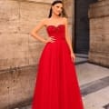 A woman exudes confidence in NC1019, a stunning strapless, floor-length red gown featuring intricate beadwork on the bodice. Her long, dark hair is styled back, and she complements her look with elegant drop earrings. She strikes a pose against the backdrop of a stone building with a closed door. Angels Formal Wear