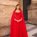 A woman stands elegantly in the vibrant red floor-length gown known as NC1019, complete with a flowing cape that enhances her grace. She accessorizes with large, dangling earrings and her hair is swept back. The backdrop features tall, ornate pillars and a stone wall, evoking an ancient or historical ambiance. Angels Formal Wear