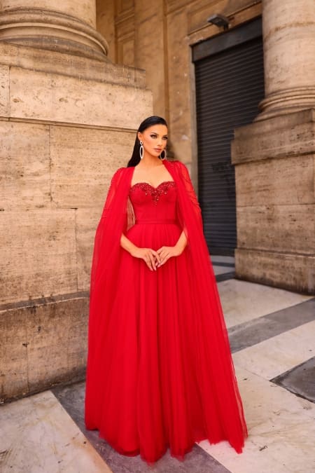 A woman stands elegantly in the vibrant red floor-length gown known as NC1019, complete with a flowing cape that enhances her grace. She accessorizes with large, dangling earrings and her hair is swept back. The backdrop features tall, ornate pillars and a stone wall, evoking an ancient or historical ambiance. Angels Formal Wear