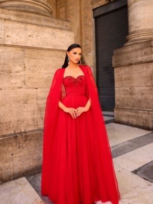 A woman stands elegantly in the vibrant red floor-length gown known as NC1019, complete with a flowing cape that enhances her grace. She accessorizes with large, dangling earrings and her hair is swept back. The backdrop features tall, ornate pillars and a stone wall, evoking an ancient or historical ambiance. Angels Formal Wear