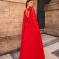 A woman in a striking gown, labeled NC1019, poses elegantly in front of a stone building. The back of her red dress features intricate beadwork and a flowing cape-like design. She wears large statement earrings, and her dark hair is styled back. The setting appears regal and historic. Angels Formal Wear