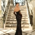 A woman stands at the bottom of a grand staircase, looking back over her shoulder. She is wearing product NC1020, a black, fitted, off-the-shoulder gown from the Dress Shop Sunshine Coast, adorned with intricate lace detailing and long, sheer gloves. The setting is elegant, with natural light streaming through large windows. Angels Formal Wear