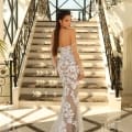 A woman in a strapless, form-fitting gown adorned with intricate lace details stands at the base of an elegant staircase. The setting, reminiscent of a Dress Shop on the Sunshine Coast, features tall windows and ornate railings, with natural light streaming in, highlighting her dress and long, flowing hair. She is wearing the stunning NC1020 gown. Angels Formal Wear