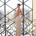 A person in the elegant SELENA NC1021 strapless silver evening gown from an Online Dress Shop stands on a staircase with intricate wrought iron railings. They are turned slightly to the side, gazing back at the camera. The background features a large geometric window with views reminiscent of the Sunshine Coast. Angels Formal Wear