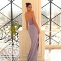 A woman in a strapless, lavender SELENA NC1021 gown stands beside a tall, white column in a sunlit room with large, grid-patterned windows. Her dress, available at our online dress shop, features a thigh-high slit and a fitted bodice with vertical sheer panels on the back. She looks over her shoulder at the camera. Angels Formal Wear