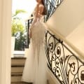 A woman in an elegant, form-fitting NC1027 white beaded gown with a tulle train stands on a staircase with intricate iron railings. She is looking back over her shoulder, and light from a large window illuminates the scene, creating an image reminiscent of Sunshine Coast school formal dresses. Angels Formal Wear