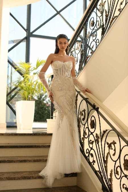 A woman stands gracefully on a staircase, leaning against the rail. She is wearing the NC1027— a strapless, fitted, white gown adorned with intricate beadwork and sheer gloves—a perfect example of school formal dresses. The background reveals large windows and a potted plant, adding elegance to the luxurious setting. Angels Formal Wear