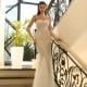 A woman stands gracefully on a staircase, leaning against the rail. She is wearing the NC1027— a strapless, fitted, white gown adorned with intricate beadwork and sheer gloves—a perfect example of school formal dresses. The background reveals large windows and a potted plant, adding elegance to the luxurious setting. Angels Formal Wear