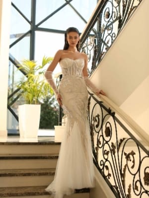 A woman stands gracefully on a staircase, leaning against the rail. She is wearing the NC1027— a strapless, fitted, white gown adorned with intricate beadwork and sheer gloves—a perfect example of school formal dresses. The background reveals large windows and a potted plant, adding elegance to the luxurious setting. Angels Formal Wear