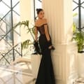 A woman in an elegant NC1028 black, off-the-shoulder gown with a fitted waist and long train stands next to a white column inside a building with tall windows. She has dark hair styled in an updo and holds black lace fabric, gazing into the distance at the epitome of formal wear for Sunshine Coast events. Angels Formal Wear