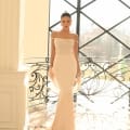 A woman stands elegantly in a sleek, off-the-shoulder NC1028 evening gown adorned with intricate beadwork on the bodice. The form-fitting dress flows into a floor-length skirt, perfect for school formal dresses. Behind her is a large window framed with black metal, allowing sunlight to illuminate the scene. Angels Formal Wear