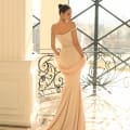 A woman in an elegant, form-fitting, off-the-shoulder light pink NC1028 gown stands in a bright, sophisticated room with large windows and intricate ironwork. Her hair is styled in a sleek updo, and she looks over her shoulder, holding a lace shawl—perfect attire for Sunshine Coast school formal dresses. Angels Formal Wear