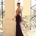 A woman in a form-fitting, off-the-shoulder NC1028 purple gown poses elegantly by a white pillar inside a building with large, geometric windows. The dress features intricate detailing on the bodice and a flowing train. Sunlight streams in, creating a warm ambiance reminiscent of the Sunshine Coast. Angels Formal Wear