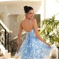 A smiling woman with her hair in an elegant bun is posing in a sunlit atrium reminiscent of the Sunshine Coast. She is wearing NC1030, a light blue, floral-patterned dress that has a laced-up back. The atrium features black wrought iron railings, marble flooring, and a potted plant in the background. Angels Formal Wear