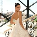 A woman with dark hair styled in an elegant updo wears NC1030, a sleeveless, white, sequined dress. She stands on a decorative staircase with ornate railings, gazing over her shoulder towards the camera. Sunlight streams in through large windows behind her, evoking the warmth of the Sunshine Coast. Angels Formal Wear