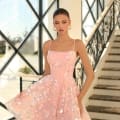 A woman stands in front of a modern windowed backdrop with stairs to her right, wearing the NC1030 sleeveless pink dress adorned with floral embroidery and sequins. She poses with one hand holding the hem of her dress and wears large hoop earrings, evoking the relaxed elegance of the Sunshine Coast. Angels Formal Wear