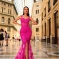 A woman stands confidently in a grand, elegant arcade, wearing the vibrant fuchsia lace CATALINA NC1031 gown with a thigh-high slit from Dress Shop Sunshine Coast. With long, dark hair and statement earrings, she poses with hands on hips under the glass domed ceiling. Afterpay is available for this stunning dress. Angels Formal Wear