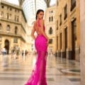 A woman in a vibrant pink CATALINA NC1031 lace gown with a long train stands in an elegant, historic indoor shopping gallery with a high glass ceiling and ornate architecture. She looks over her shoulder at the camera, showcasing the back details of her dress from Dress Shop Sunshine Coast. Angels Formal Wear