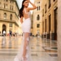 A woman in the elegant CATALINA NC1031 sheer white lace gown, available at Dress Shop Sunshine Coast, stands in an ornate, sunlit indoor courtyard. She poses with one hand on her head and looks over her shoulder. The courtyard features intricate architectural details, arched ceilings, and a marble floor, with people visible in the background. Angels Formal Wear