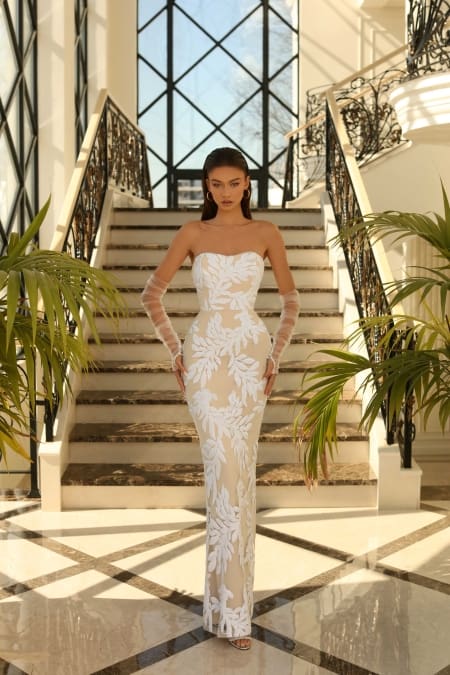 A woman elegantly draped in the NC1032, a strapless, floor-length dress adorned with white floral patterns, stands before a grand staircase featuring wrought iron railings. The bright and airy Sunshine Coast atmosphere is reflected in the stairs and large windows behind her. She complements her look with sheer gloves. Angels Formal Wear