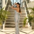 A woman stands on a grand staircase, striking a pose with one hand on her head while wearing the dazzling NC1032—a form-fitting, strapless gown adorned with intricate silver sequins. She completes the look with long, sheer gloves. The setting features large windows and ornate railings, accentuated by nearby potted plants, perfectly encapsulating the elegance of the Sunshine Coast. Angels Formal Wear