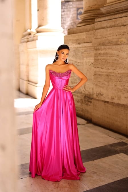 A woman stands in an elegant stone corridor, wearing a strapless, bright pink satin gown named NC1033. The gown features beaded detailing at the neckline. Her dark hair is styled neatly, and she accessorizes with large hoop earrings. Light filters through the checkered floor, highlighting her stunning ensemble. Angels Formal Wear