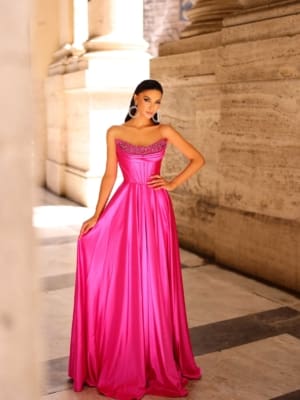 A woman stands in an elegant stone corridor, wearing a strapless, bright pink satin gown named NC1033. The gown features beaded detailing at the neckline. Her dark hair is styled neatly, and she accessorizes with large hoop earrings. Light filters through the checkered floor, highlighting her stunning ensemble. Angels Formal Wear