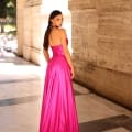 A woman with long, dark hair stands on a marble floor next to a stone wall, wearing a strapless hot pink gown. She looks over her shoulder at the camera, and sunlight streams in from the left, highlighting the architectural details around her in an almost cinematic glow akin to the NC1033. Angels Formal Wear