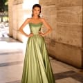 Clad in an exquisite green satin gown featuring an off-the-shoulder design and a beautifully embellished bodice, a person poses elegantly. They stand confidently in the grand, sunlit stone corridor marked NC1033, their hair styled back and adorned with long earrings that enhance their graceful aura. Angels Formal Wear