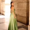 A woman in the NC1033 strapless, floor-length green satin gown stands in a corridor with tall columns and stone walls. She looks over her shoulder towards the camera, with sunlight highlighting her dress and hair. Angels Formal Wear