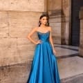 A woman poses against a stone wall, wearing the NC1033 strapless blue satin gown adorned with embellishments on the bodice. She accessorizes with long earrings and accents her stance with her hand on her hip. The background showcases textured stone columns, contributing to the architectural setting. Angels Formal Wear