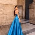 A woman is standing on a marble floor, wearing an elegant, strapless, floor-length royal blue gown. She has long, dark hair and is facing backwards, looking over her shoulder. The background features a tall stone column and an arched doorway marked with the product name NC1033. Angels Formal Wear