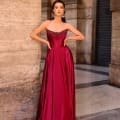 A woman stands confidently in the NC1033, a strapless, floor-length burgundy dress featuring intricate detailing on the bodice. She poses with hands on hips against a backdrop of textured stone walls and a marble floor, exuding elegance and poise. Angels Formal Wear