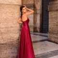 A woman in a strapless, flowing dark red gown stands against a stone wall, with one hand touching her long, dark hair. Sporting large, elegant earrings, she exudes grace as the NC1033 glimmers faintly. The background features a column and an archway. Angels Formal Wear