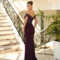 A woman stands on a marble floor at the base of a grand staircase with ornate black railings. Dressed in an elegant AVA NC1035, off-shoulder, dark purple evening gown and with her hair styled in an updo, she basks in the sunlight filtering in, illuminating the luxurious setting. Angels Formal Wear