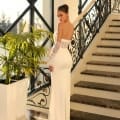 A woman stands on a marble floor at the base of an ornate staircase. She is wearing the ASHLEY NC1036, a strapless white gown with long lace gloves, her back turned to the camera. Natural light streams through large windows, illuminating a potted plant beside her in this stunning setting reminiscent of an Sunshine Coast online dress shop. Angels Formal Wear