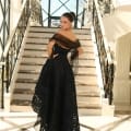 A woman stands at the bottom of an elegant staircase on the Sunshine Coast, wearing the NC1037, a strapless black floor-length gown featuring an intricate lace pattern. Her hair is styled in an updo, and she drapes a sheer black shawl over her shoulders. The setting is bright with large windows and ornate iron railings. Angels Formal Wear