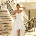 A woman wearing the elegant NC1037 off-shoulder white lace dress poses on the steps of a grand staircase adorned with intricate wrought iron railings. The high-low hem of her dress beautifully showcases her white heels. Bathed in sunlight, the scene evokes the feel of a perfect day on Sunshine Coast. Angels Formal Wear