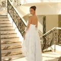 A woman wearing the NC1037, a strapless white gown adorned with an intricate lace pattern, stands at the bottom of an elegant staircase. The staircase, reminiscent of Sunshine Coast luxury, features ornate black railings and marble steps. She looks over her shoulder back towards the camera as sunlight streams in. Angels Formal Wear