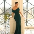 A woman in a dark green, off-the-shoulder MISCHA NC1040 gown stands beside a tall white column, gazing back over her shoulder. Her hair is styled in an elegant updo, and she wears matching NC1040 earrings. The background features large windows with a geometric grid pattern and indoor plants. Angels Formal Wear