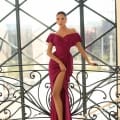 A woman in a deep red off-the-shoulder MISCHA NC1040 dress with a high slit stands poised against a decorative railing. She is in front of a large, geometric window structure, allowing ample natural light to highlight her elegant attire. She wears strappy high heels and carries an air of confidence. Angels Formal Wear