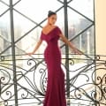 Wearing the MISCHA NC1040 gown in magenta, a person stands gracefully on a polished marble floor beside an intricate black wrought-iron railing. Sunlight streams through expansive geometric windows, casting a luxurious and airy glow over the elegant scene. Angels Formal Wear