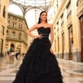 A woman stands confidently in an elegant, strapless MONTANA NC1047 black ruffled gown within a grand, ornate indoor setting with a high, arched glass ceiling. She has one hand on her hip and is smiling. The architecture features intricate detailing and tall columns, reminiscent of MONTANA's historic charm. Angels Formal Wear