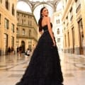 A woman in an elegant MONTANA NC1047 black gown poses in a grand, ornate indoor gallery with a glass dome ceiling. The polished marble floor reflects the classical architectural details of the walls. Other visitors can be seen walking in the background, adding to the serene ambiance reminiscent of Montana's natural beauty. Angels Formal Wear