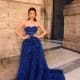 A woman stands confidently in front of a rustic stone wall and large door, wearing the elegant MONTANA NC1047 strapless blue ball gown with layers of tulle. With her hands on her hips and statement earrings, she exudes a chic, sophisticated look that captures Montana's timeless grace. Angels Formal Wear