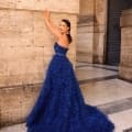 A woman stands against a large stone column, wearing the elegant MONTANA NC1047 gown—a strapless royal blue ruffled dress that flows onto the floor. Her dark hair is pulled back, and she accessorizes with dangling earrings. The background features a tall, closed dark door and beige stone walls, capturing the timeless elegance reminiscent of the MONTANA NC1047. Angels Formal Wear