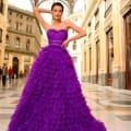 A woman in a strapless, tiered purple gown poses confidently in an ornate, domed indoor architectural setting. The background features grand archways and large windows, with the MONTANA NC1047 model adding an extra touch of elegance to the scene. A few people are visible in the distance. Angels Formal Wear