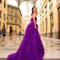 A woman in a ruffled, bright purple gown poses gracefully on marble steps in a grand, ornate indoor setting with a high glass ceiling. She looks over her shoulder towards the camera, exuding the timeless charm of MONTANA NC1047, with people walking in the background. Angels Formal Wear